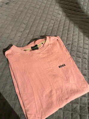 Rosa t-shirt från BOSS - Säljer en stilren rosa t-shirt från BOSS med diskret logga på bröstet. T-shirten har rund halsringning och är tillverkad i mjuk bomull med stretch för extra komfort. Perfekt till jeans eller shorts för en clean look.