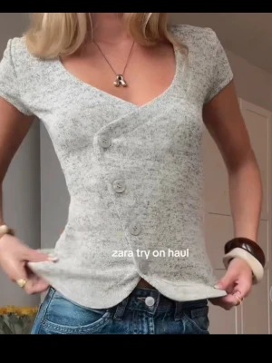 Ljusgrå omlottopp från Zara - Kortärmad ljusgrå omlottopp från Zara med v-ringning och fyra dekorativa knappar framtill. Toppen har en lätt melerad look och är tillverkad i mjukt, stretchigt material som sitter snyggt på kroppen. Perfekt till jeans eller kjol för en trendig vardagsstil. Den har kvar lappen. Helt ny. 