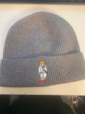 Grå  mössa Polo Bear - Grå ribbstickad mössa från Polo Ralph Lauren med Polo Bear-brodyr framtill där björnen har astronautdräkt. Klassisk uppvikt kant och mjukt material som håller dig varm. Perfekt accessoar för att lyfta din streetwear-look.