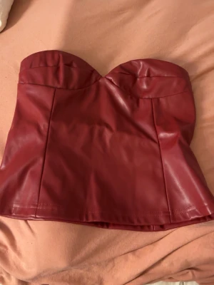 Röd axelbandslös skinnkorsett - Snygg röd axelbandslös korsettopp i skinnimitation. Toppen har en bandeauformad överdel och markerade sömmar framtill.