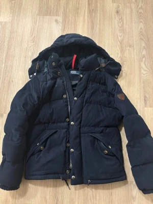 Mörkblå vinterjacka från Polo Ralph Lauren i Manchester - Snygg mörkblå dunjacka från Polo Ralph Lauren med avtagbar huva, dragkedja och knappar framtill.  Jackan har två stora fickor med lock och detaljer i brunt läder på ärmen. Perfekt för kalla dagar och riktigt skön passform. Storleken är XS men sittter mer som S.