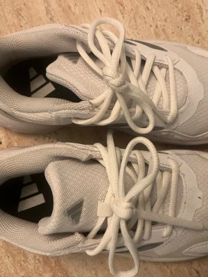 Vita Adidas sneakers med silvriga detaljer - Fräscha vita Adidas sneakers med silvriga ränder på sidorna och klassisk snörning. Ovandelen är i mesh och syntet för en sportig look. Sulan har tydlig struktur och märkningen Cloudfoam och Adiwear syns på sidan.