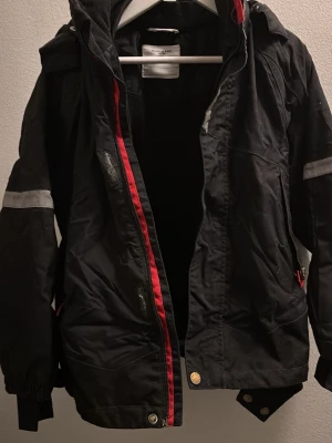 Svart vinterjacka från P.O.P/Polarn O. Pyret barn 122/128 - Svart vinterjacka från P.O.P  med huva och röda detaljer längs dragkedjan. Jackan har reflexband på ärmarna, flera fickor och muddar vid ärmsluten. 