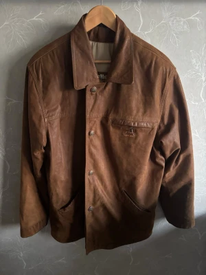 Brun skinnjacka från Camel Active - Snygg brun skinnjacka från Camel Active med klassisk krage och tryckknappar framtill. Jackan har flera fickor, inklusive en bröstficka med logga, och är helfodrad i beige tyg. Perfekt för dig som gillar tidlös och avslappnad stil.