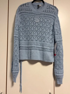 Ljusblå virkad stickad tröja H&M -  Ljusblå stickad tröja från H&M med virkat hålmönster och snörningsdetalj i sidan. Tröjan har långa ärmar, ribbade muddar  