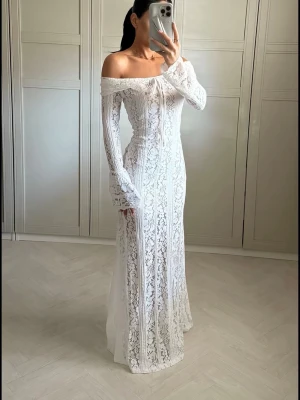 Vit off-shoulder spetsklänning - Elegant lång vit klänning med off-shoulder design och långa ärmar. Klänningen är helt täckt av vacker spets och har en figurnära passform som ger en romantisk och stilren look. Perfekt för speciella tillfällen.