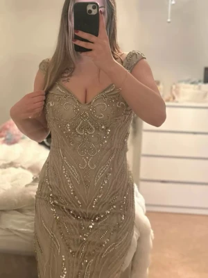Champagnefärgad långklänning med paljetter - Säljer en elegant champagnefärgad långklänning med glittriga paljettdetaljer och pärlor. Klänningen har v-ringning, smala axelband och figurnära passform. Perfekt för bal eller festliga tillfällen där du vill glänsa extra mycket.