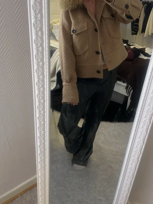 Beige jacka med fuskpäls från Meotine - Kort beige jacka från Meotine med stor krage i fuskpäls. Jackan har två stora bröstfickor med lock och svarta knappar framtill. Materialet är strukturerat och fodret är mörkgrönt. Perfekt för dig som vill ha en trendig och mysig look.