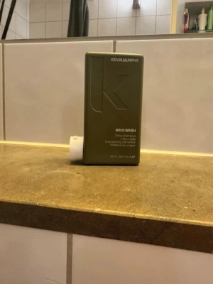 Kevin Murphy MAXI.WASH detoxschampo - Grön rektangulär flaska med Kevin Murphy MAXI.WASH detoxschampo. Schampot är speciellt framtaget för att rengöra håret på djupet och bevara färg. Flaskan har en minimalistisk design med präglad logga och vit skruvkork.