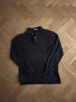 Mörkblå långärmad piké från Polo Ralph Lauren - Klassisk mörkblå långärmad pikétröja från Polo Ralph Lauren med röd broderad logga på bröstet. Tröjan har krage, knäppning med två knappar och ribbade muddar vid ärmsluten. Perfekt för en stilren och avslappnad look.