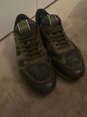 Valentino Rockrunner camo sneakers - Säljer ett par Valentino Rockrunner sneakers med camouflage-mönster i gröna och bruna toner. Skorna har detaljer i mocka och skinn, samt klassisk snörning. Ikonisk Valentino-logga på plösen och cool kontrastdetalj vid hälen. Dom nöts lite i hälen i skon men ingenting som syns/känns och dom är riktigt snygga nu till sommaren! Pris kan diskuteras vid snabb affär!