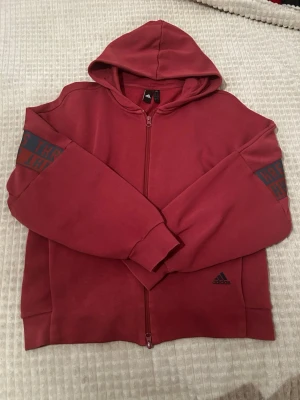 Vinröd hoodie från Adidas - Snygg vinröd hoodie från Adidas med dragkedja och huva. Adidas-logga på framsidan och tryck på ärmarna. Tillverkad i mjukt material som känns skönt mot huden. Perfekt för en avslappnad och sportig stil.