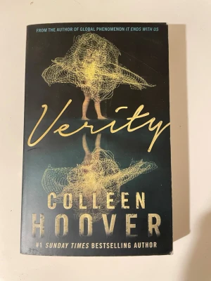 Verity av Colleen Hoover - En spännande bok med titeln 'Verity' av Colleen Hoover. Omslaget är svart med guldfärgad text och ett konstnärligt motiv av en kvinna i gult. Boken är i pocketformat och har en modern, stilren design. Perfekt för dig som gillar nervkittlande berättelser.