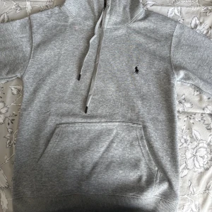 Grå hoodie från Polo Ralph Lauren (tar emot byten) - Snygg ljusgrå hoodie från Polo Ralph Lauren med klassisk broderad logga på bröstet. Tröjan har huva med snörning, känguruficka och ribbade muddar. Tillverkad i mjuk bomull för en skön och avslappnad look.