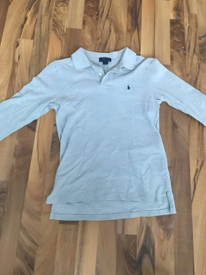 Ljusgrå pikétröja från Ralph Lauren - Klassisk ljusgrå pikétröja från Ralph Lauren med krage, knappar vid halsen och den ikoniska broderade loggan på bröstet. Tröjan har lång ärm och är tillverkad i mjuk bomull som känns skön mot huden. Perfekt för en stilren och avslappnad look.