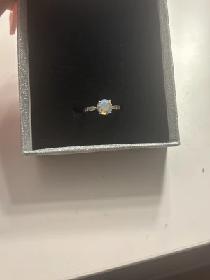 Silverring med regnbågsskimrande sten - Snygg silverfärgad ring med en rund, regnb sopal-liknande sten i mitten. Ringen har små gnistrande detaljer på sidorna av stenen och ger ett elegant och trendigt intryck. Perfekt för dig som gillar unika smycken med lite extra bling.st av 16c