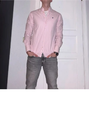 Ljusrosa skjorta med knappar - Säljer en stilren ljusrosa skjorta med klassisk krage och knäppning framtill. Skjortan har lång ärm och en liten broderad logga på bröstet. Perfekt för dig som vill ha en enkel men snygg skjorta till din outfit.