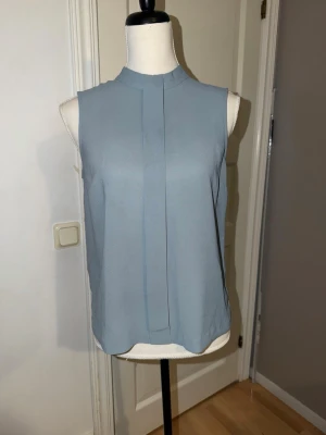 Ljusblå ärmlös blus med hög krage - Stockh LM  - Snygg ljusblå blus med hög krage och ärmlös design. Blusen har en rak passform och ett stilrent veck framtill. Perfekt att matcha med både jeans och kjol för en clean look.