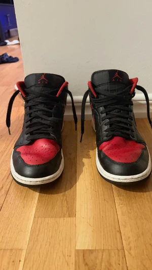 Nike Air Jordan 1 svart/röd sneakers - Säljer ett par klassiska Nike Air Jordan 1 sneakers i svart och rött skinn med vita sulor. Ikonisk design med snörning, rund tå och Air Jordan-logga på sidan. Perfekta för dig som gillar streetwear och vill sticka ut med en tidlös modell.