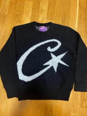 Corteiz Sweater Svart - Säljer en svart stickad tröja från Corteiz i storlek L. Tröjan har ett stort vitt stjärnmotiv på framsidan och en liten broderad logga på ärmen. Klassisk rund halsringning och ribbade muddar. Perfekt för dig som gillar grafiska detaljer. Passar även bra till M.