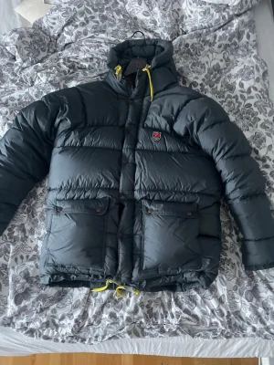 dunjacka från Fjällräven  - Säljer enriktigt fluffig dunjacka från Fjällräven. Jackan har stora fickor framtill, gul snörning nertill och klassisk Fjällräven-logga på bröstet. Yttertyget är i polyamid och fyllningen består av 90% gåsdun och 10% fjäder. Perfekt för kalla dagar!
