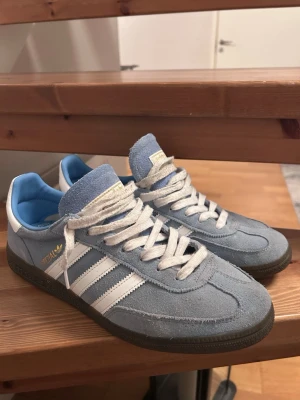 Adidas Spezial ljusblå sneakers mocka - Säljer ett par Adidas Spezial sneakers i ljusblå mocka med vita detaljer och klassiska tre ränder på sidan. Skorna har vit snörning, vit hälkappa och gummisula. Guldtexten 'SPEZIAL' syns på sidan och innersulan är blå med Adidas-logga. Orginalsnören och box tillkommer. Pris går att diskutera och hör av dig vid minsta fundering😊