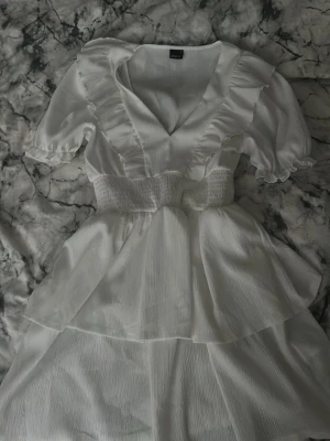 Vit volangklänning med v-ringning - Söt vit klänning med v-ringning, volangdetaljer och korta puffärmar. Midjan är smockad för en bekväm passform och kjolen har ett lager med volang. Perfekt för sommarens alla tillfällen.