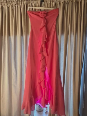 Rosa långklänning från Adoore - Supersnygg rosa långklänning från Adoore med volangdetalj framtill och en söt rosett upptill. Klänningen är axelbandslös och har dragkedja i ryggen. Perfekt för dig som vill sticka ut med en feminin och elegant look. Använd endast en gång på balen och tvättad två gånger. Katt finns i hemmet!