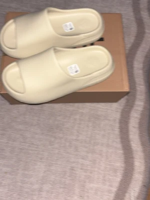 Beige Yeezy Slide tofflor - Säljer ett par stilrena beigea Yeezy Slide tofflor från Adidas. De har en minimalistisk design med öppen tå och platt sula, tillverkade i syntetmaterial för en mjuk och bekväm känsla. Perfekta för chill dagar hemma eller när du vill ha en clean look.