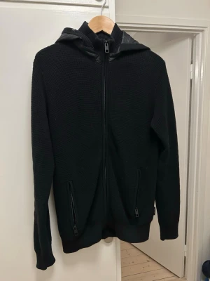 Cardigan jacka  - Svart cardigan jacka med hel dragkedja framtill och två fickor med dragkedja. Stickad struktur och huva med tygdetaljer. Perfekt för en avslappnad och stilren look. Materialet är mjukt och bekvämt, passar till många outfits. Luvan går att ta av o på