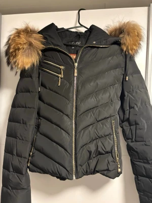 Svart dunjacka med fuskpäls från Hollies - Svart dunjacka från Hollies med snygga guldfärgade dragkedjor och detaljer. Jackan har en huva med fuskpäls i bruna toner och flera praktiska fickor med dragkedja. Perfekt för kalla dagar och ger en cool, urban look.