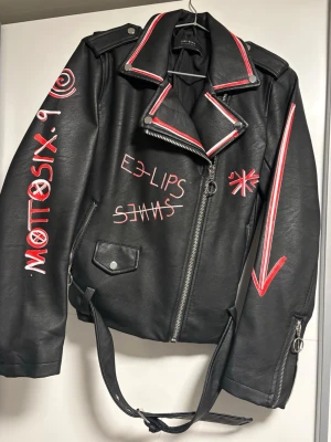 Svart skinnjacka med röda detaljer Zara - Cool svart skinnjacka från Zara Basic med röda och vita grafiska detaljer, text och pilar. Jackan har klassisk bikerkrage, dragkedja och bälte i midjan. Perfekt för dig som vill sticka ut med en edgy stil.