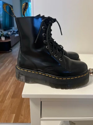 Svarta Dr. Martens kängor i skinn - Säljer ett par klassiska svarta Dr. Martens kängor i skinn med gul söm runt sulan och grov platt sula. Kängorna har snörning framtill och den ikoniska flärpen bak. Perfekta för dig som vill ha en edgy och tidlös stil.