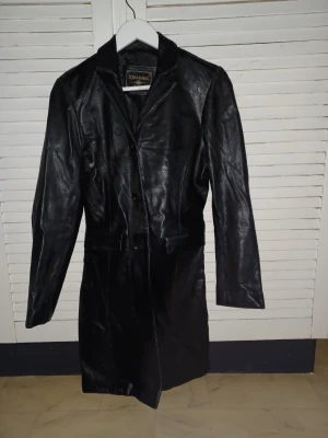 Svart skinnrock från Tomahawk - Snygg svart skinnrock från Tomahawk med klassisk krage och knäppning framtill. Rocken har en rak passform och är tillverkad i äkta skinn, vilket ger en cool och tidlös look. Perfekt för dig som vill sticka ut med en edgy stil.