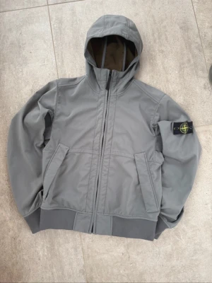 Grå vindjacka från Stone Island - Snygg grå vindjacka från Stone Islande med huva och ikonisk patch på ärmen. Jackan har dragkedja framtill, två snedställda fickor och ribbade muddar vid ärmslut och midja. Perfekt för dig som vill ha en stilren och funktionell jacka, säljer för att den är för liten.