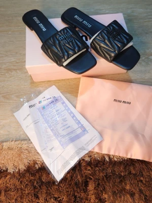 Svarta sandaler  - Snygga svarta sandaler quiltad ovandel i skinn och broderad logga. Sandalerna har fyrkantig tå och platt sula, perfekt för en trendig look. Levereras med originalkartong och dustbag.