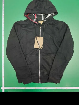 Burrbery kofta - Svart Burberry-kofta med huva och dragkedja. Klassisk och stilren modell som passar till vardags. Använd men i bra skick, inga större skador eller hål. Bekväm och varm, perfekt för höst och vinter.