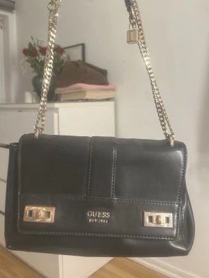 Svart axelväska från Guess - Snygg svart axelväska från Guess med guldfärgade kedjor och detaljer.. Tillverkad i skinnimitation och har dekorativa sömmar samt vridlås i guld på framsidan. Den är väl använd därav priset. Köptes för ca 1200kr förra året. Väskan har repor som man kan se på bilden. Kedjan är även lite missfärgad. Jätte fin väska annars och bra i storlek mycket får plats.