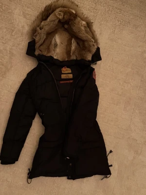 Svart dunjacka med fuskpäls från Fjällräven - Svart dunjacka från Fjällräven med lång passform och huva fodrad med mjuk fuskpäls. Jackan har dragkedja framtill, justerbar midja och klassisk Fjällräven-logga på ärmen. Perfekt för kalla vinterdagar.