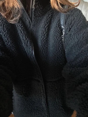 Svart teddyjacka - Mysig svart teddyjacka med dragkedja framtill och hög krage. Jackan har en oversized passform och är tillverkad i fluffigt teddy-material.