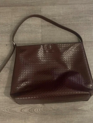 Vinröd axelväska i flätad skinnimitation - Snygg vinröd axelväska med flätad struktur och bred axelrem. Väskan har en stilren, rektangulär form och är tillverkad i skinnimitation. Perfekt för dig som gillar klassisk men trendig design.