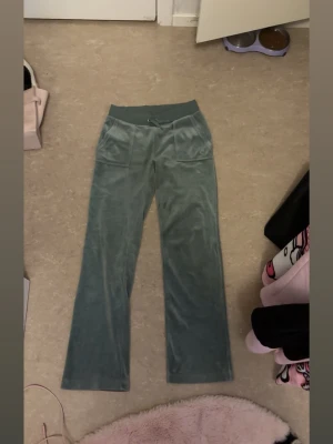 Gröna mjukisbyxor i velour - Mysiga gröna mjukisbyxor i velour med bred resårmidja och snörning. Byxorna har raka ben och två fickor fram samt två bakfickor med knappdetaljer. Perfekta för chill dagar eller när du vill vara extra bekväm.