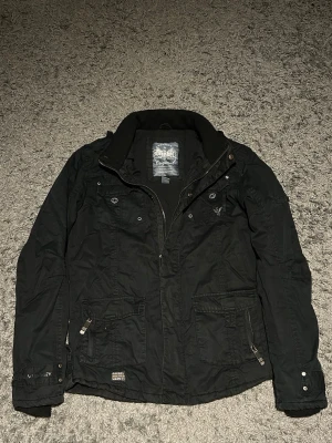 Svart vindjacka från diesel - Svart vindjacka från diesel med flera fickor, dragkedja och knappdetaljer framtill. Jackan har hög krage och justerbara muddar. Perfekt för blåsiga dagar och har en stilren look med diskret logga på bröstet.