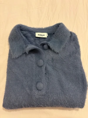 Blå fluffig kofta från Dobber - Mysig och fluffig blå kofta från Dobber med korta ärmar och stora tygklädda knappar framtill. Koftan har klassisk krage och är gjord i ett mjukt, lurvigt material som känns extra skönt mot huden. Perfekt för dig som gillar retrovibbar och vill sticka ut.