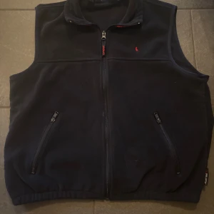 Polo Ralph Lauren väst  - Svart fleeceväst från Polo Ralph Lauren med röd broderad logga på bröstet. Västen har två fickor med dragkedja, hel dragkedja framtill och en hög krage. Perfekt för lager-på-lager och chill stil. Aldrig använd 