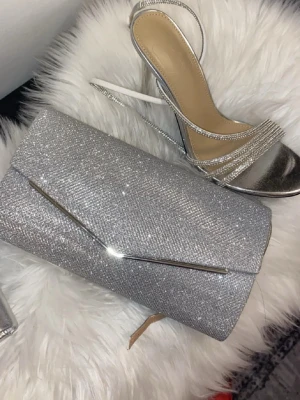 Bal accessoarer paket  - Snygg kuvertväska i glittrig silver med stilren metall-detalj på framsidan. Perfekt storlek för mobilen och det viktigaste. Väskan har en elegant och modern look som lyfter din outfit till nästa nivå.