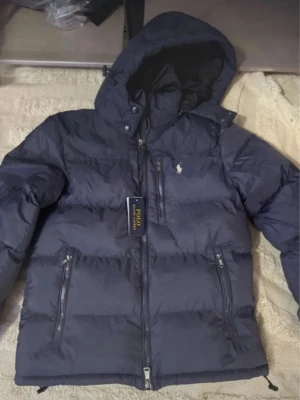 Mörkblå dunjacka Polo Ralph Lauren - Snygg mörkblå dunjacka från Polo Ralph Lauren med huva och dragkedja framtill. Jackan har två sidofickor med dragkedja och klassisk Polo-logga på bröstet. Perfekt för kalla dagar med sin puffiga design och stilrena look.