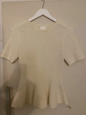 Beige stickad topp med volangkant - Superfin beige stickad topp med kort ärm och rund hals. Modellen är figurnära upptill och har en snygg volangkant nedtill som ger en feminin siluett. Perfekt att styla med jeans eller kjol för en trendig look.