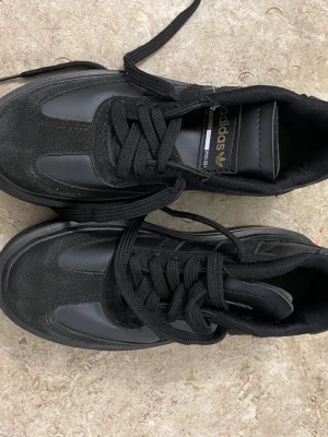 Svarta Adidas sneakers med plattform - Säljer ett par svarta Adidas sneakers med platt sula och snygg plattform. Skorna har klassiska tre ränder på sidan, gulddetaljer på häl och plös samt snörning. Materialmix av mocka och syntet ger en cool look. Perfekt för dig som gillar streetstyle och vill sticka ut.