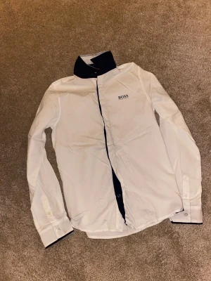 Vit skjorta från Hugo Boss slim fit - Vit långärmad skjorta från Hugo Boss med marinblå detaljer på krage och knappslå. Slim fit-modell med klassisk krage och knappar framtill. Boss-logga broderad på bröstet. Perfekt för en clean och stilren look.
